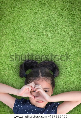 Happy smiling girl kid lying on green grass lawn looking through heart love shape hand hole: National children's eye health and safety month in August Child's eyesight healthcare awareness CSR concept