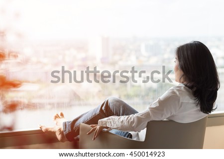 Business woman lifestyle at home sitting on modern chair in living room looking out of window toward beautiful cityscape downtown urban river landscape city life w/ sunlight effect: Easy happy people