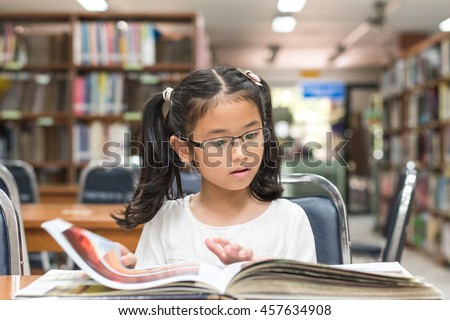 Smart little asian child girl w/ eyeglasses reading book school background: Lovely cute young female student kid opening flipping book in archive resource collection room: National library lover month