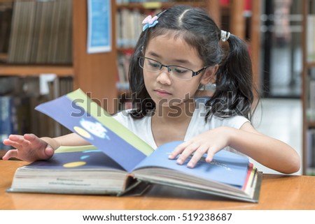 Happy little asian child girl wear eyeglasses reading book school background: Lovely cute young student kid opening flipping book in archive resource collection room: National library lover month week