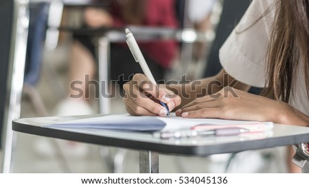 Student hand holding pen writing doing examination with blurred abstract background university students in uniform attending exam classroom educational school: Blurry view college people in class room