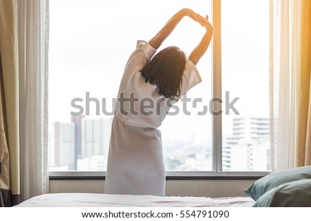 Happy healthy woman back view waking up stretching in bed room hotel/ home interior at glass wall window, city landscape background: Simple lifestyle people in cozy indoor comfortable relaxing room