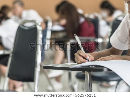 Student hand holding pen writing doing examination with blurred abstract background university students in uniform attending exam classroom educational school: Blurry view college people in class room