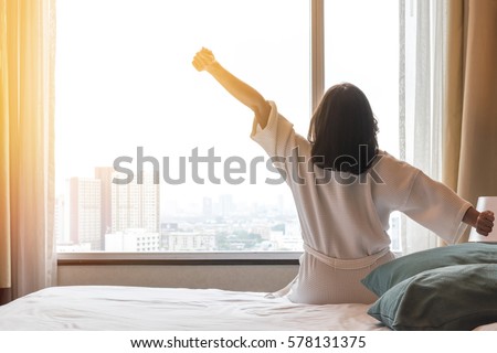 Happy healthy woman back view waking up stretching in bed room hotel/ home interior at glass wall window, city landscape background: Simple lifestyle people in cozy indoor comfortable relaxing space