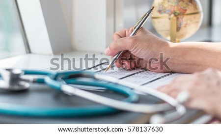 Doctor\'s hand working using pen writing on medical prescription blank paper form for patients with stethoscope on table desk: Physician taking note on empty RX paperwork in hospital/ clinic interior