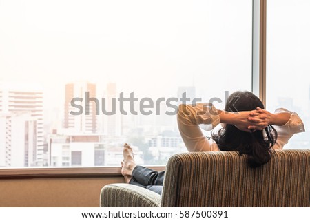Business woman lifestyle at home sitting on cozy modern chair living room looking out of window toward beautiful cityscape downtown urban landscape city life, warm sunlight effect: Easy happy people