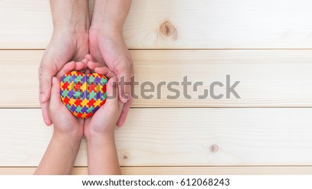 Red love heart in puzzle jigsaw pattern on child kid palms supported by mother's parent caregiver hand on light white pine wood background symbolic campaign for World Autism Awareness day on April 2th