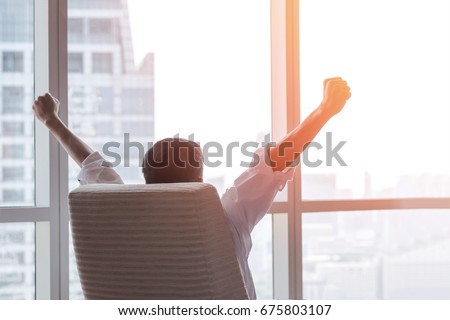 Business achievement concept with happy businessman relaxing in office room, resting and raising fists with ambition looking forward to city building urban scene through glass window