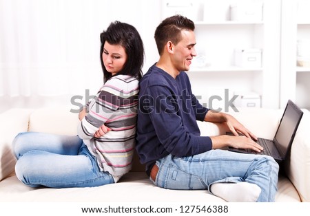 Young couple sitting back to back on the sofa, man enjoying the computer, woman is sad and feeling neglected