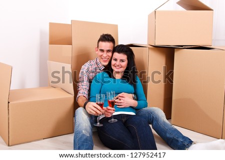 Happy couple on the moving day sitting on the floor near cardboard boxes, drinking wine, having break