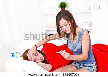Beautiful sick girl lying in bed, her friend takes care of her, measuring her temperature