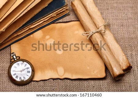 Above view of old pocket watch with vintage paper rolls and aged books. Burlap background.