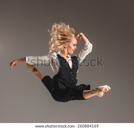 Beauty blond woman in a black suit and white shirt in ballet jump on a gray background