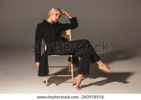 Beauty blond woman  in a black coat on chair on a gray background