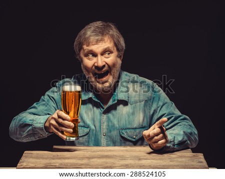 The front view of handsome screaming man as fan in denim shirt with glass of beer, sitting at the wooden table. Concept of emotional fan, interested showing something
