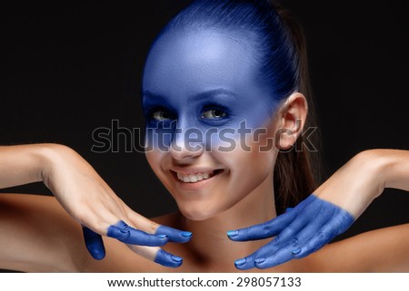 Portrait of a smiling young woman who is posing covered with blue paint in the studio on a black background. girl\'s hands around the face and painted blue