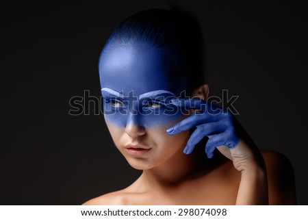 Portrait of a young woman who is posing covered with blue paint in the studio on a black background. girl\'s hand around the face and painted blue