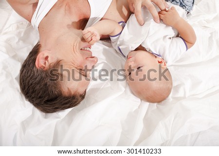 Young smiling father with his nine months old son  lying upside down on the bed at home on white home background