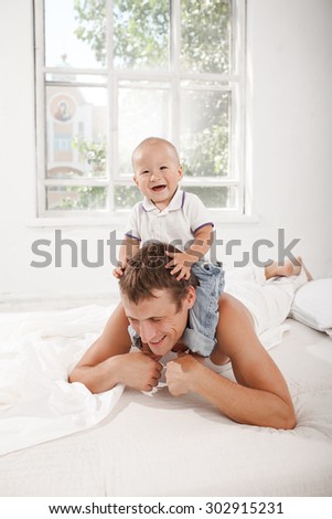 Young smiling father with his nine months old son on the bed at home on white home background. son sitting on his father's shoulders