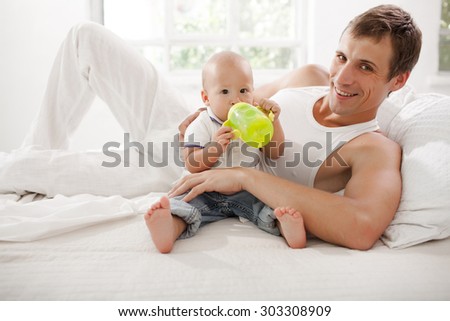 Young smiling father with his nine months old son on the bed at home on white home background. Child drinking water