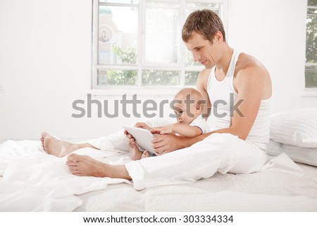 Young father with his nine months old son sitting on the bed at home on white home background with digital tablet