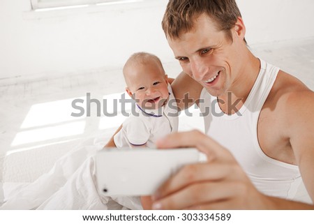 Young smiling father with his nine months old son on the bed at home on white home background. dad taking selfie photo with telephone camera