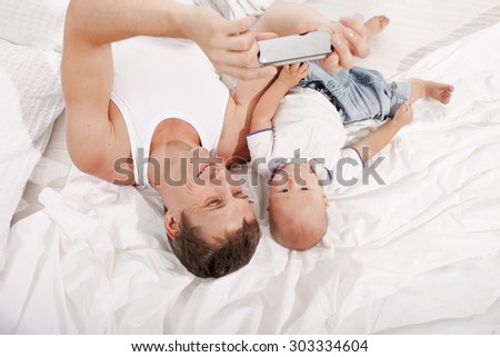 Young smiling father with his nine months old son  lying upside down on the bed at home on white home background. dad taking selfie photo with telephone camera