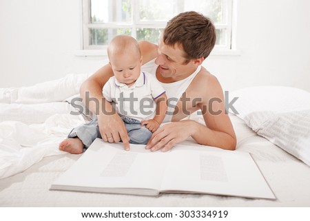 Father and baby sitting together and reading book on the bed at home on white home background