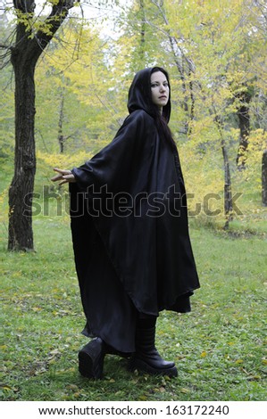 Black hooded dryad woman in the autumn forest, turning back at you while walking as if beckoning