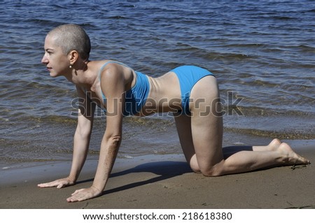 Bald woman in yoga position marjaryasana-bitilasana, or cat-cow pose, on a beach.