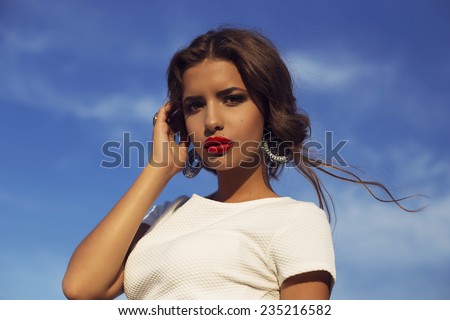 Outdoor fashion photo of-the beautiful Italian woman brunette with curly hair in a white blouse and black skirt and jevelry with bright makeup on a sky background enjoying the fresh air