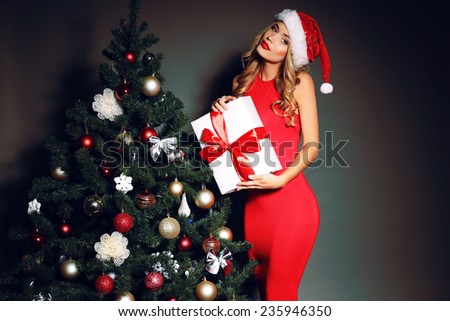 Christmas photo of sexy woman with curly blond hair in a red Santa-hat and fitting red dress wtith white smile  holding a gift-boxes near Christmas tree