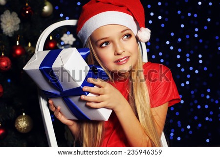 cute little girl whith long blond hair and blue beautiful eyes in santa hat and red suit surprised of Christmas present on the backgroud of holiday shining lights