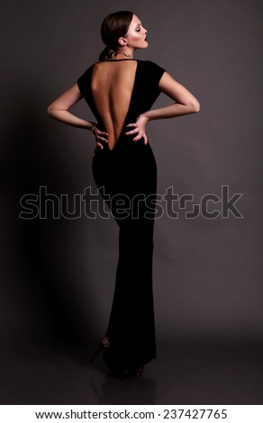 Elegant fashion photo of glamor beautiful brown-haired woman in a black sexy dress with open back wearing gold jewelry and posing in the studio,luxury