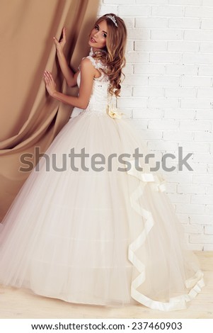 festive wedding photo of chic sexy bride in white evening lace dress with diadem like a princess with blond beautiful curly hair posing in the studio,smiling with white teeth
