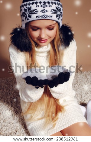 Fashion photo of cute little girl with long blond hair wearing a hat with ornament and bubonic,gloves, white sweater and knitted stockings sitting on the carpet,smiling,surprise and  catches a snow