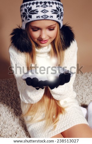 Fashion photo of cute little girl with long blond hair wearing a hat with ornament and bubonic,gloves, white sweater and knitted stockings holding a snow and sitting on the carpet,magic