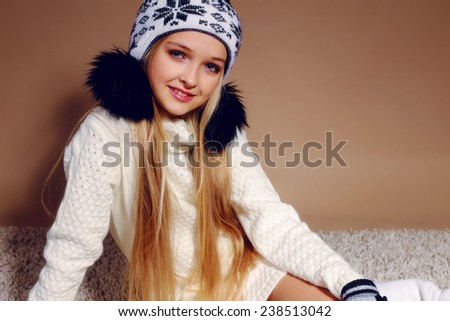 Fashion photo of cute little girl with long blond hair wearing a hat with ornament and bubonic,gloves, white sweater and knitted stockings sitting on the carpet and smiling