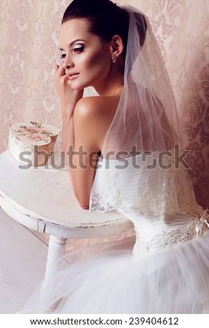 festive wedding photo of chic sexy bride in white evening dress with lace corset and bridal veil, with brunette hair, sitting at the table in bedroom
