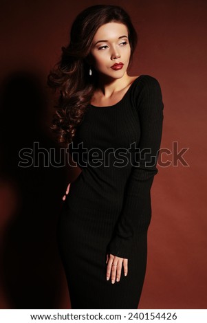 fashion photo of sexy brunette woman in black dress and heeled shoes with red lips and curly hair posing in studio