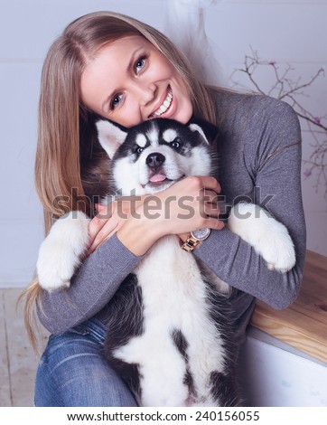 Studio photo of beautiful blond girl with long straight hair,big smile, white teeth wearing in gray sweater, jeans and holding a cute little puppy of Husky