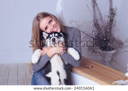 Studio photo of beautiful blond girl with long straight hair,big smile, white teeth wearing in gray sweater, jeans and holding a cute little puppy of Husky
