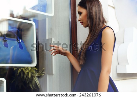 fashion outdoor photo of beautiful elegant lady with dark long hair a wearing blue dress standing at the shop window and selecting a bag on sale