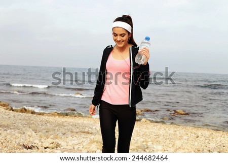 sport outdoor photo of beautiful young brunette woman in black sport suit with headband smiling and listening to music on headphones by the sea