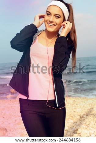 sport outdoor photo of beautiful young brunette woman in black duffle with headband smiling and listening to music on headphones by the sea
