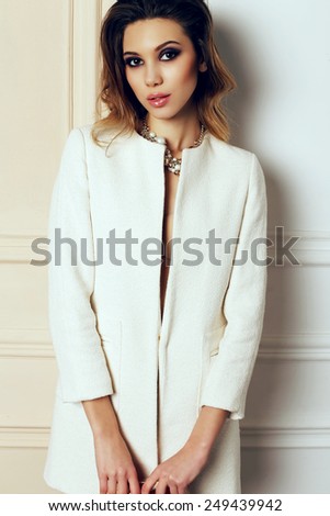 fashion photo of sexy brunette woman in elegant white coat and gold necklace with volume hair and bright evening makeup, posing at studio with classic interior