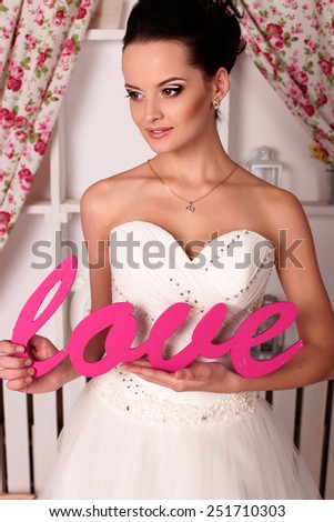 Valentine\'s Day photo of chic sexy bride in white evening dress with brunette hair posing at home near the cabinet, holding a wood pink word of love