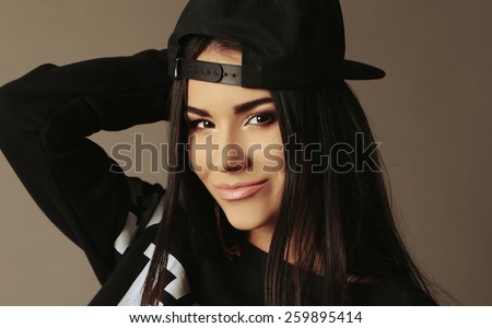 Fashion sportive photo of sexy tanned Asian woman with dark long hair bright makeup, wearing black sweater,cap posing at studio