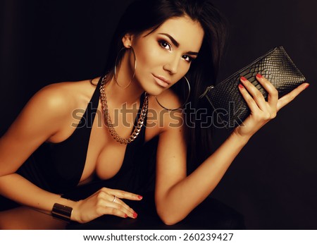 Fashion portrait of sexy tanned Asian lady with dark long hair wearing black lingerie and beautiful gold jewelry, holding a leather clutch and posing at studio