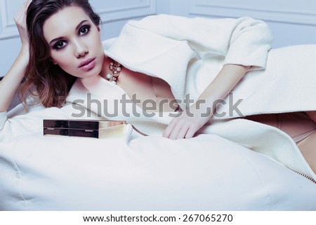 fashion photo of sexy brunette woman in elegant white coat and gold necklace with volume hair and bright evening makeup posing at studio with classic interior